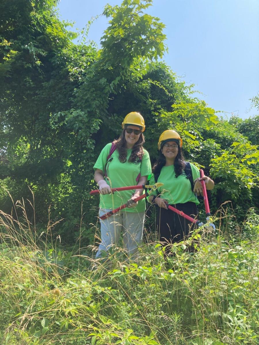 Emily and Stacey, Conservation interns, pose in the bushes with shears. They are both smiling and wearing green Town T-shirts and yellow hard hats.